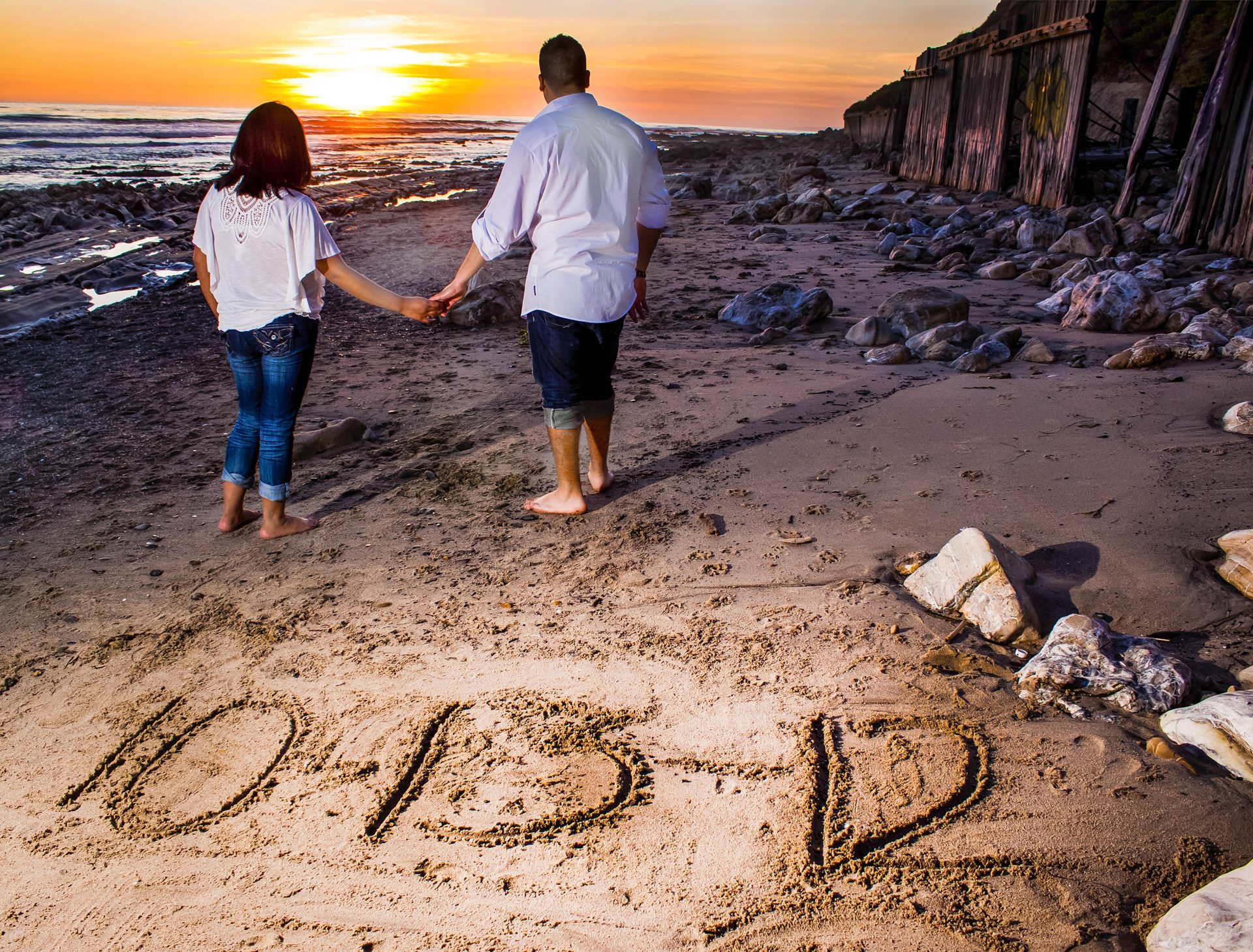 Couple walking hand in hand toward a beach sunset, their date written in the sand