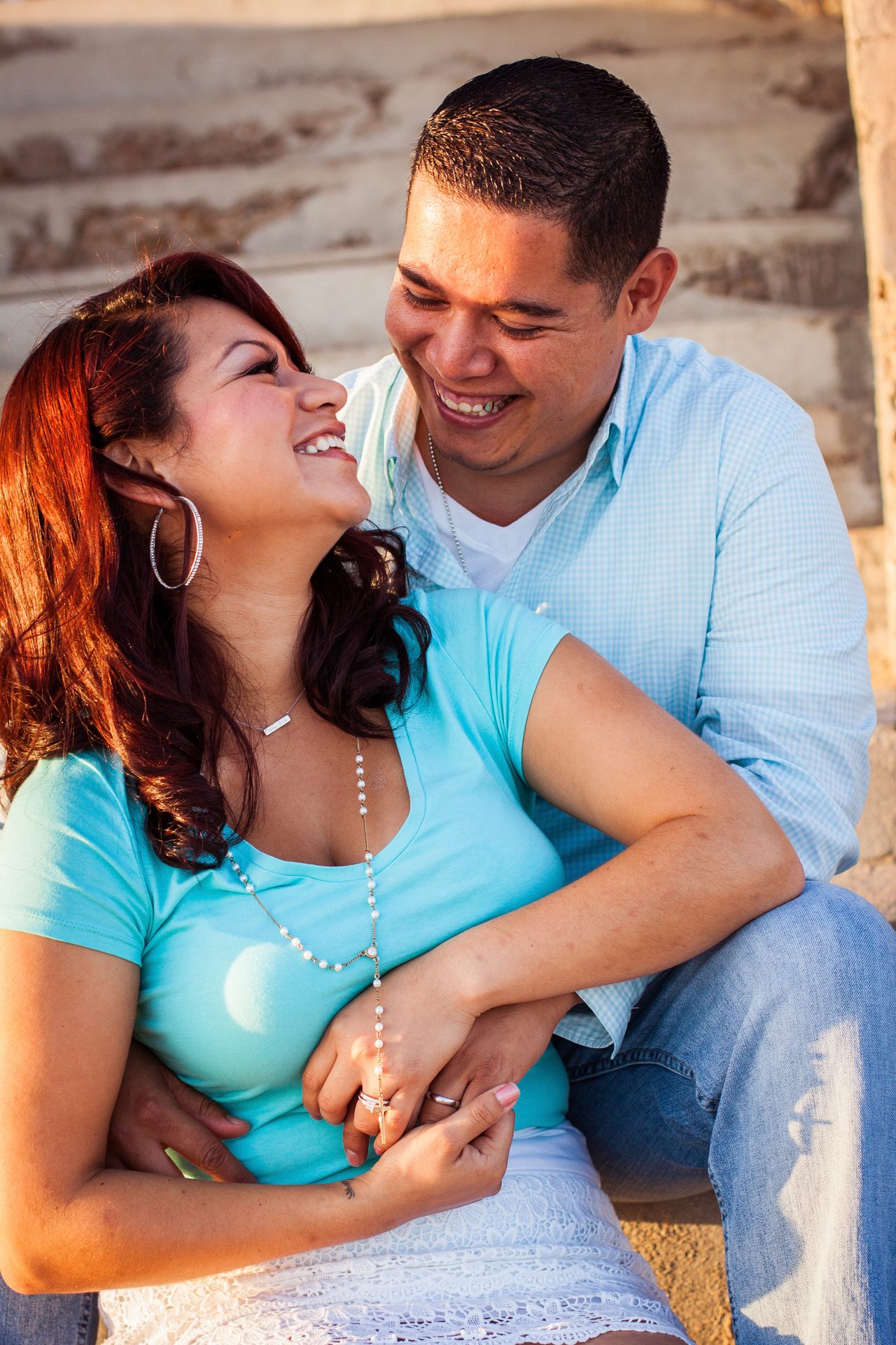 Engaged couple laughing together on sunlit steps, his arms wrapped around her