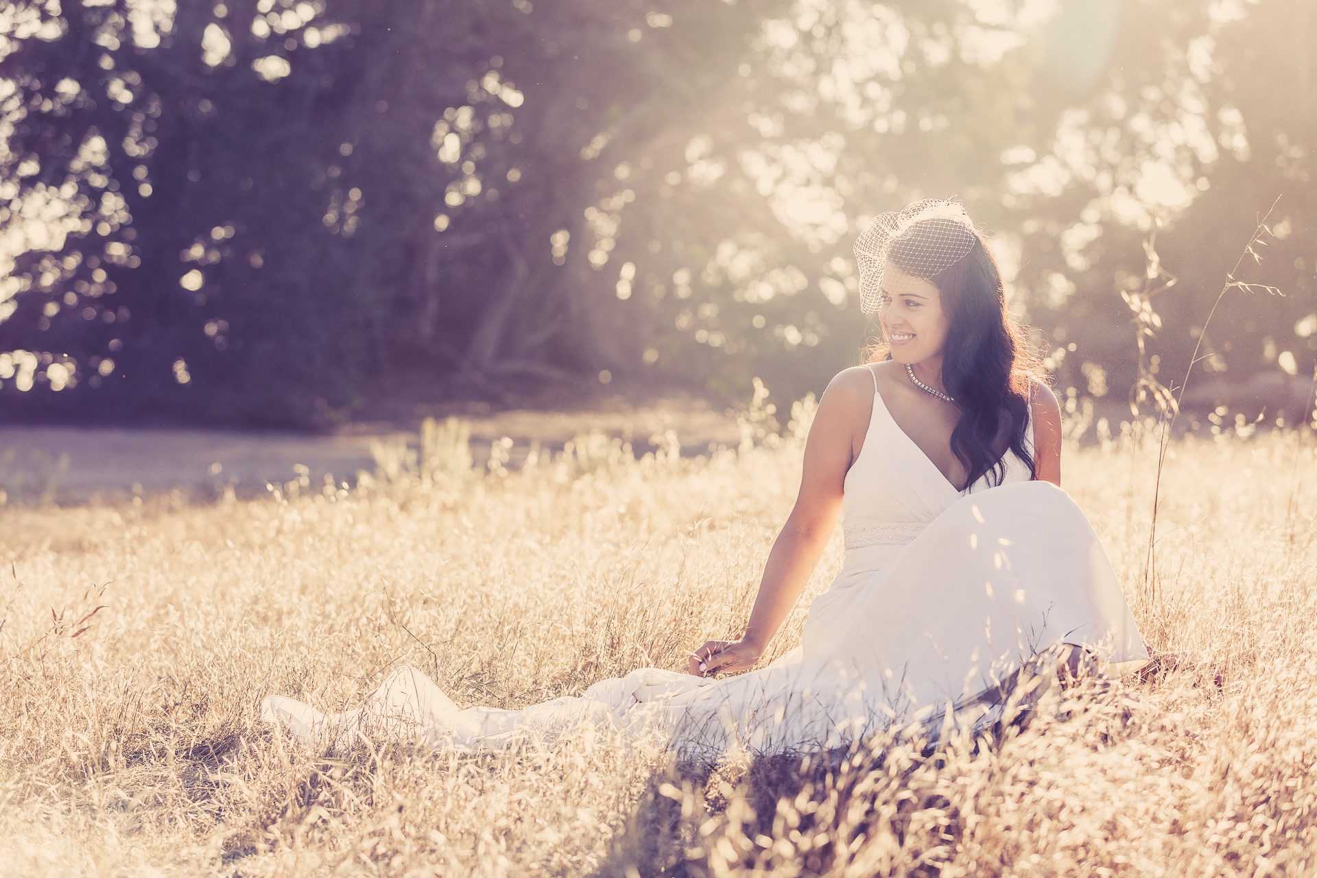 Woman in a white dress sitting in golden dry grass as the sun flares through the trees