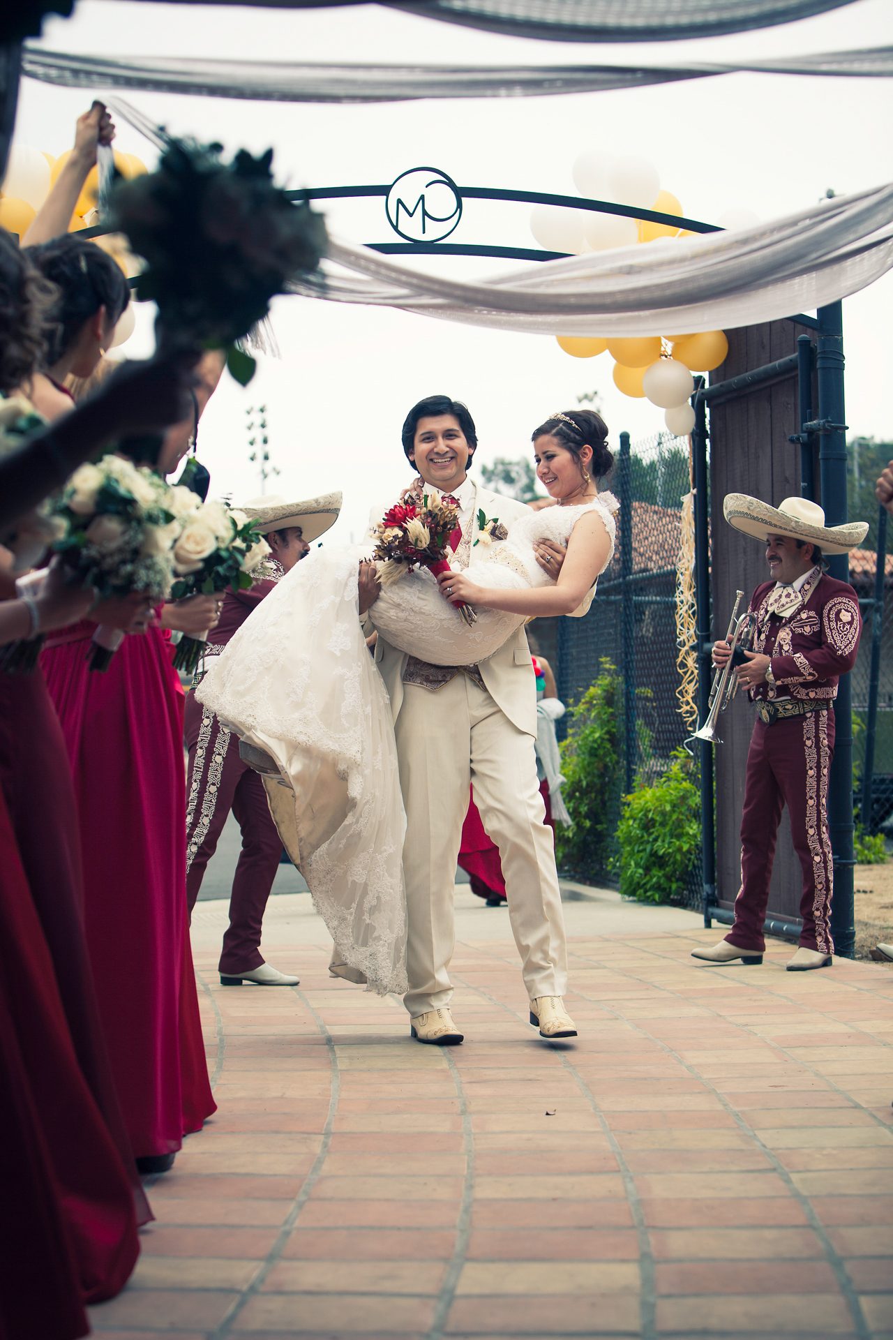 Man carrying a woman in white beneath a flower arch as mariachi musicians play