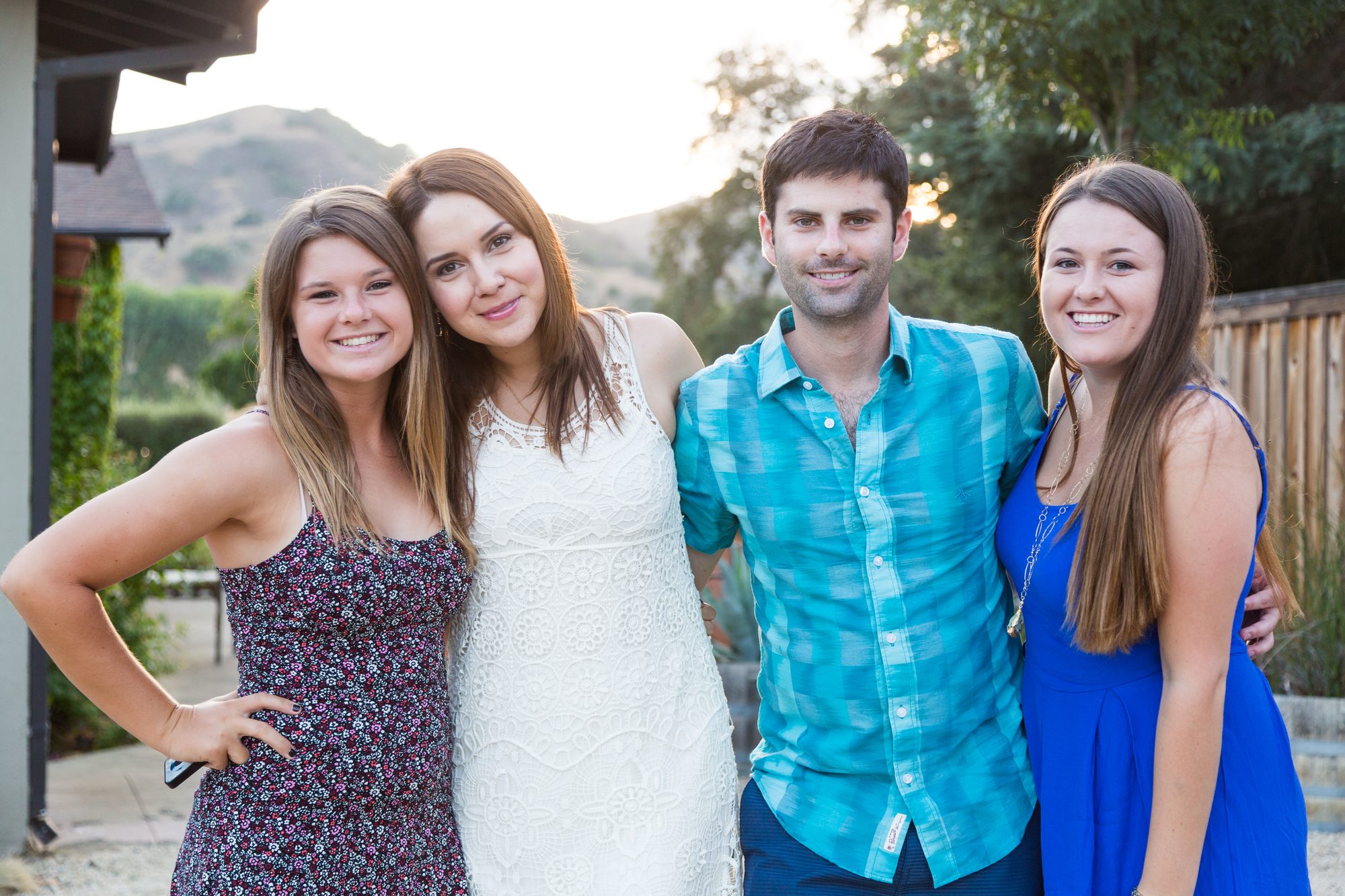 Four young adults standing arm in arm in a backyard at golden hour, hills behind