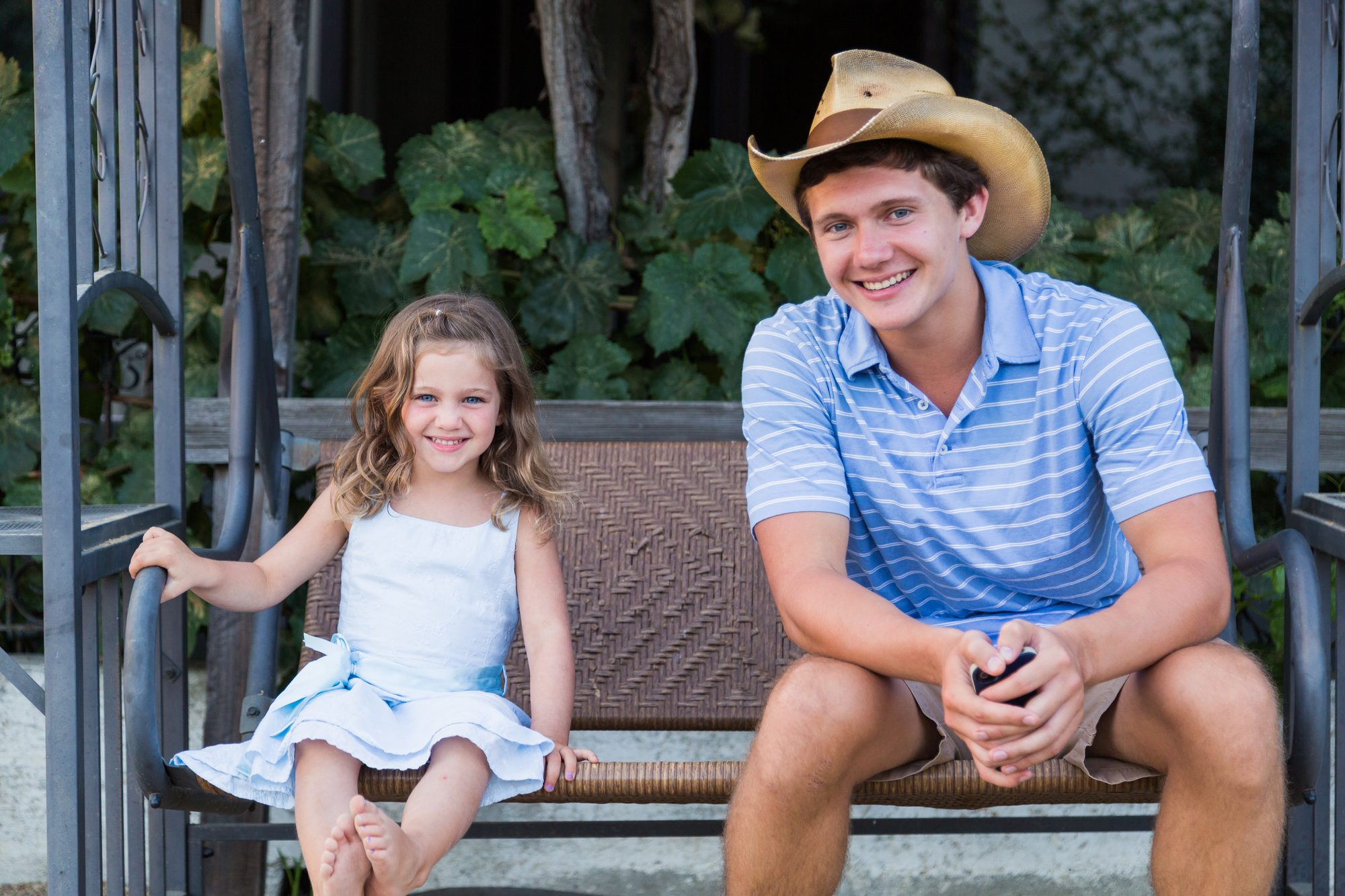 Little girl in a light-blue dress and a teenager in a cowboy hat sitting on a porch swing