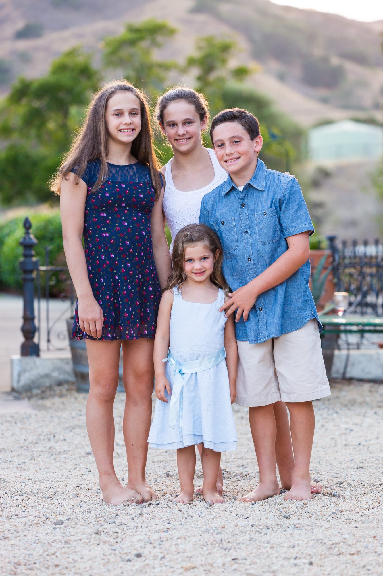Four barefoot kids standing together on gravel with golden hills behind them