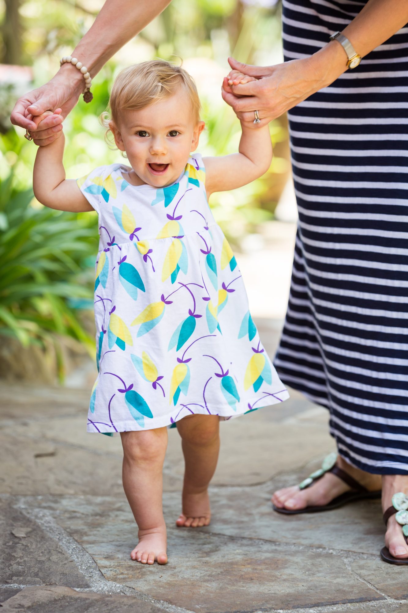 Blonde toddler in a floral dress taking steps on a flagstone path while holding grown-ups' hands