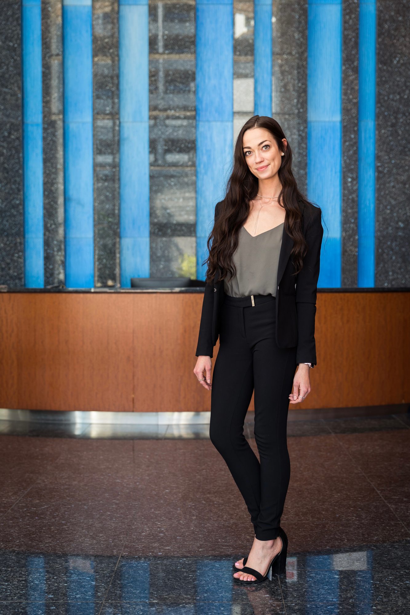 Full-length portrait of a woman with long wavy brown hair in a black blazer, olive top and black pants standing in a building lobby, a blue and granite wall behind her