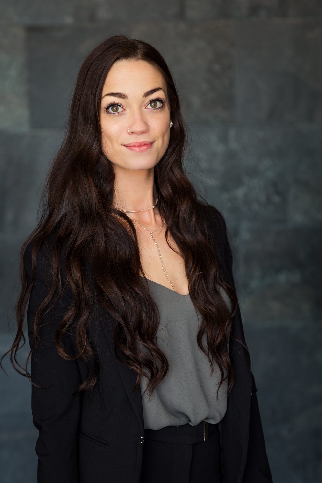 Headshot of a woman with long wavy brown hair in a black blazer against a dark slate wall