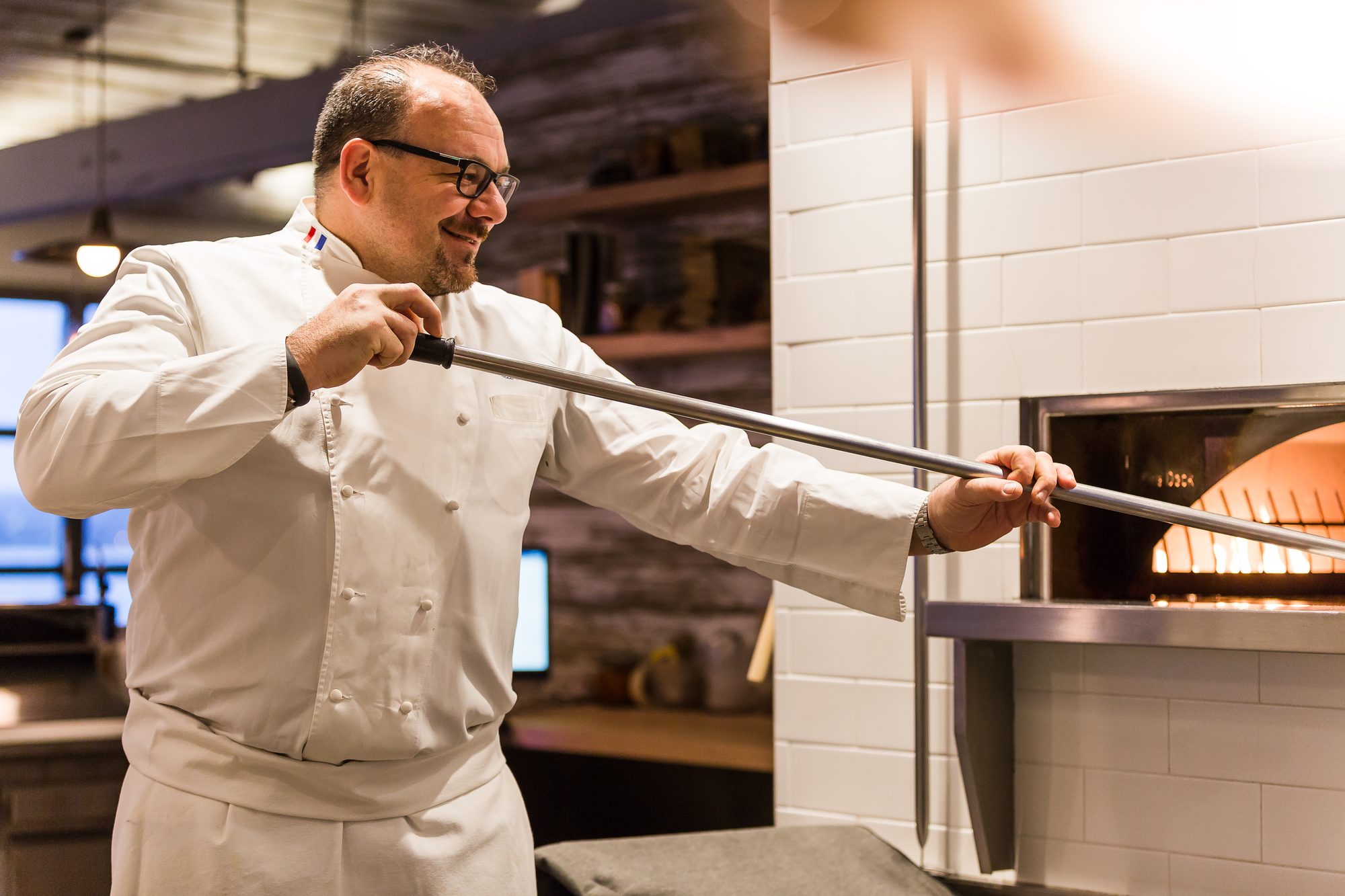 Chef in whites working a long peel at a wood-fired oven in a restaurant kitchen