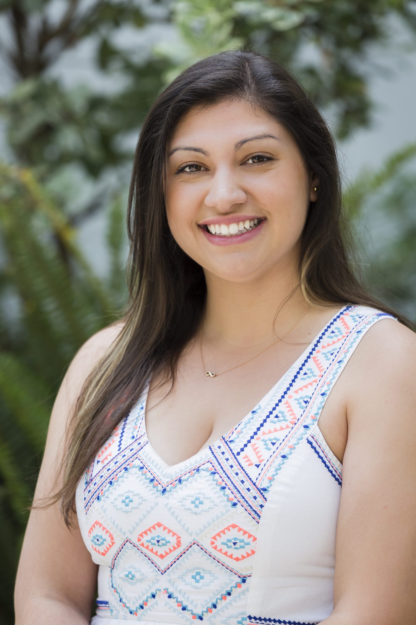 Smiling woman with long dark hair in an embroidered white top, greenery behind