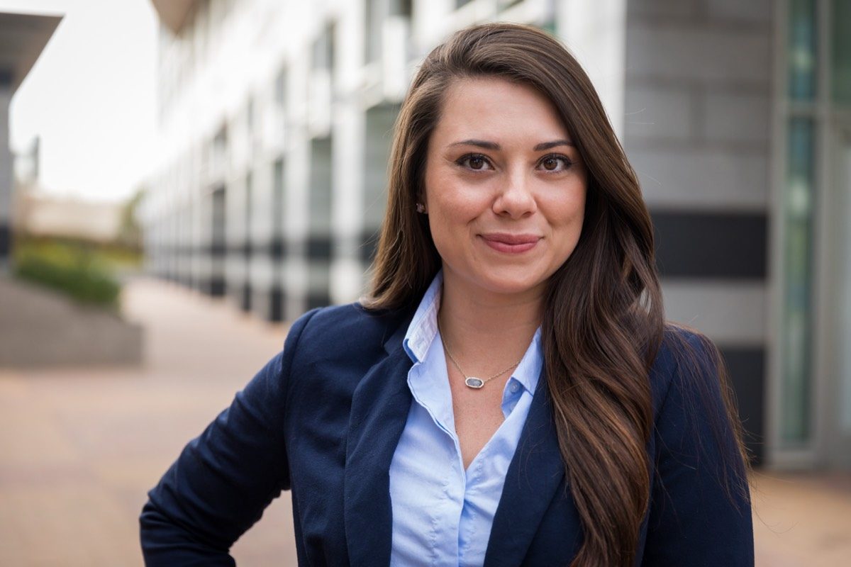 Smiling woman with long brown hair in a navy blazer outdoors, modern building behind
