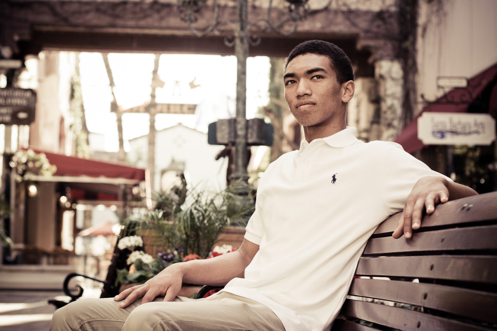 High school senior in a white polo seated on a wooden bench in a sunlit plaza