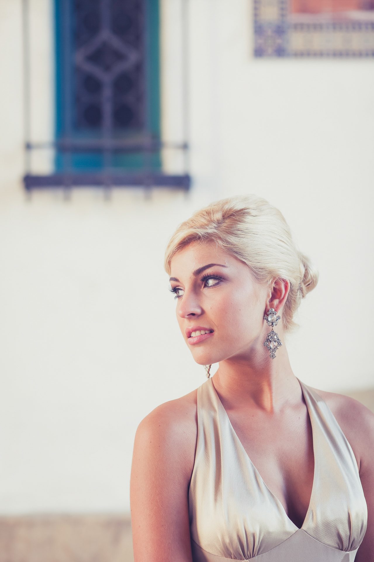Woman with platinum hair in a champagne satin halter dress and chandelier earrings, gazing off beside a white Spanish-style wall